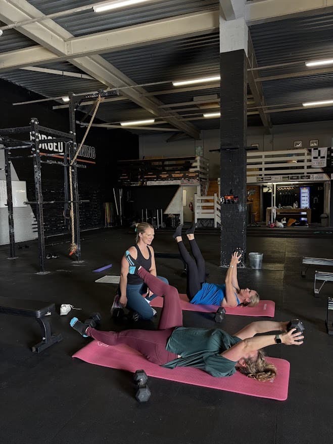Kate kneeling on the gym floor coaching two clients through dumbbell work on mats